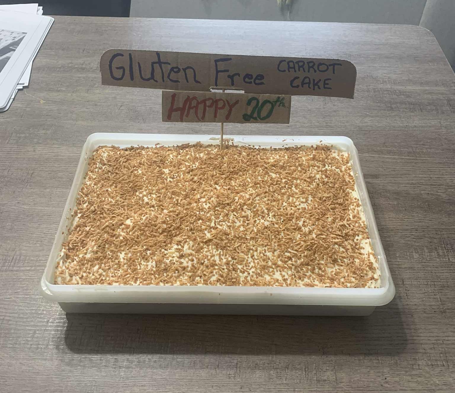 A picture of a carrot cake with icing and coconut and sign that says Gluten free - happy 20th